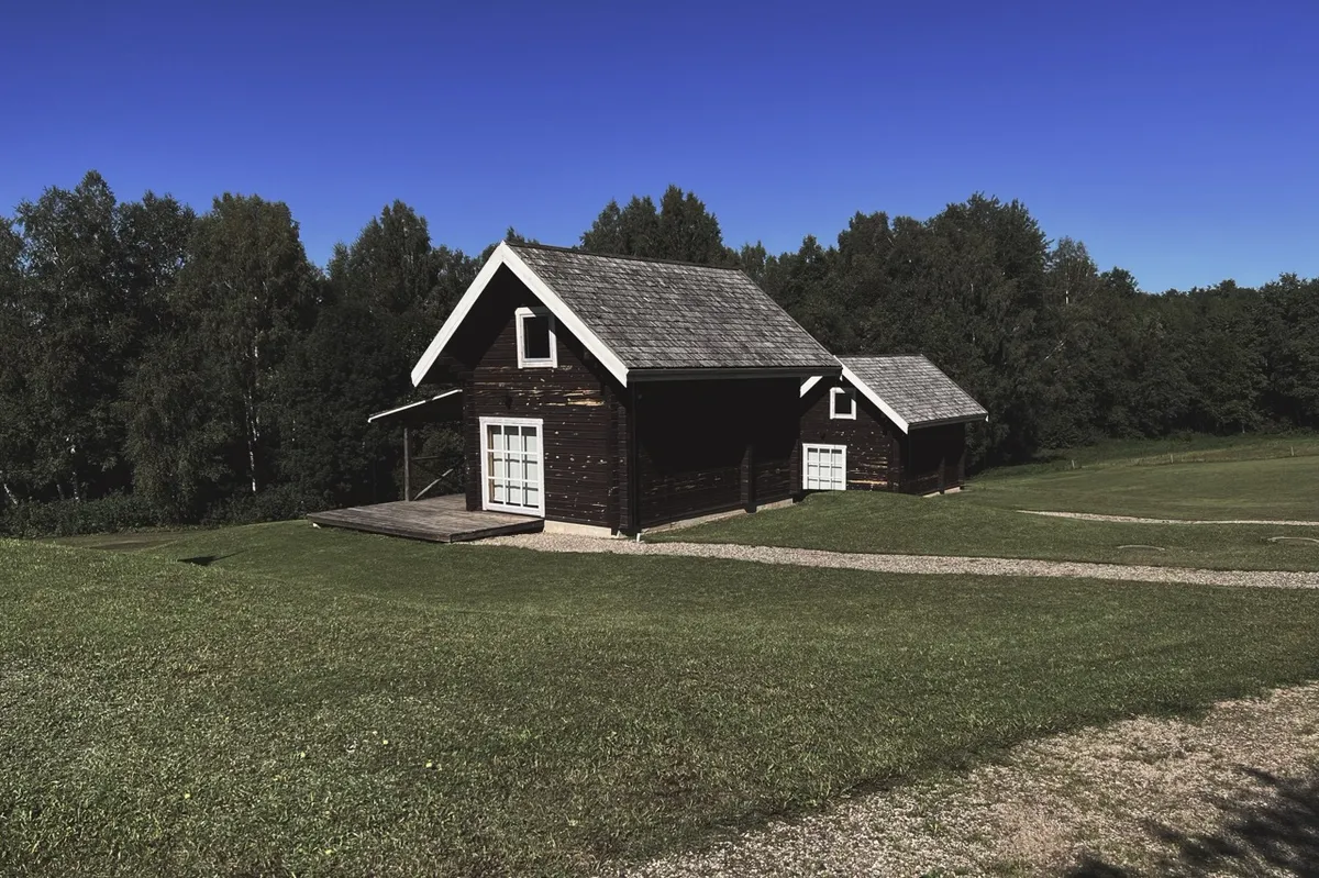 A rustic wooden cabin stands on a lush green lawn, surrounded by a forest under a clear blue sky, with a gravel path leading to its entrance.