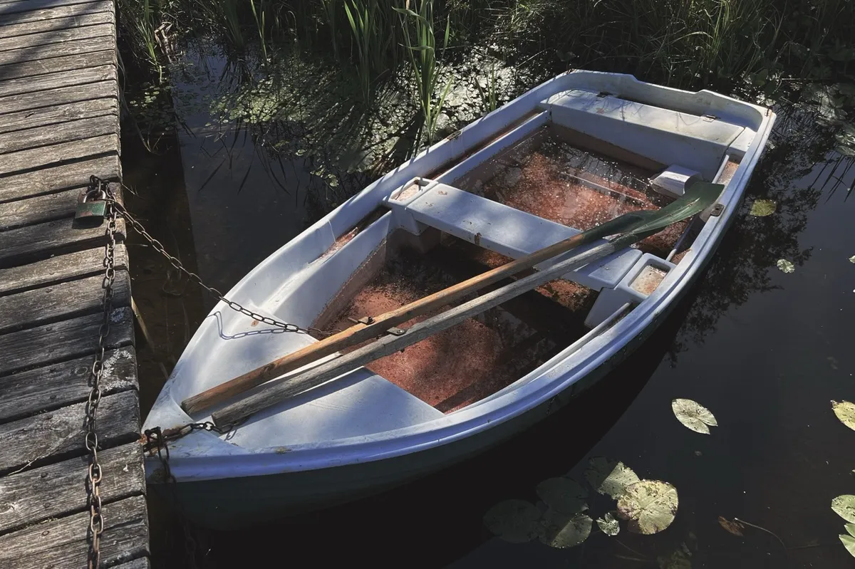A small rowboat, with oars resting inside, is moored to a wooden dock by a chain. It floats among lily pads and reeds in calm water.