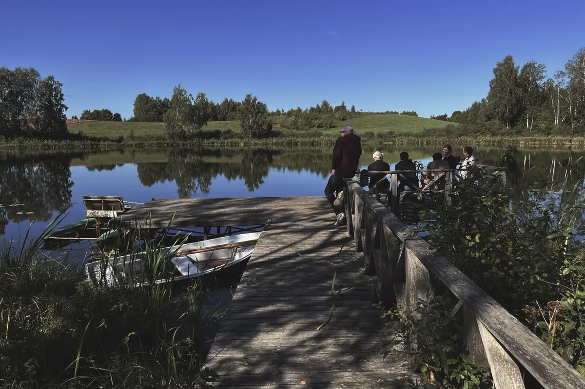 A wooden dock extends over a calm lake, with boats moored alongside. People sit and stand on the dock, surrounded by lush green trees under a clear blue sky.