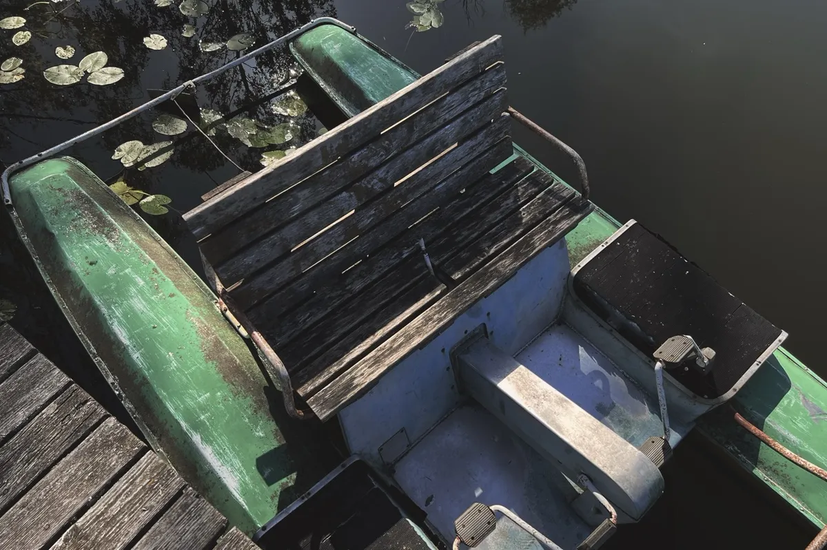 Wooden paddle boat with worn green paint and a bench seat, floating beside a wooden dock surrounded by water lilies in a calm pond.