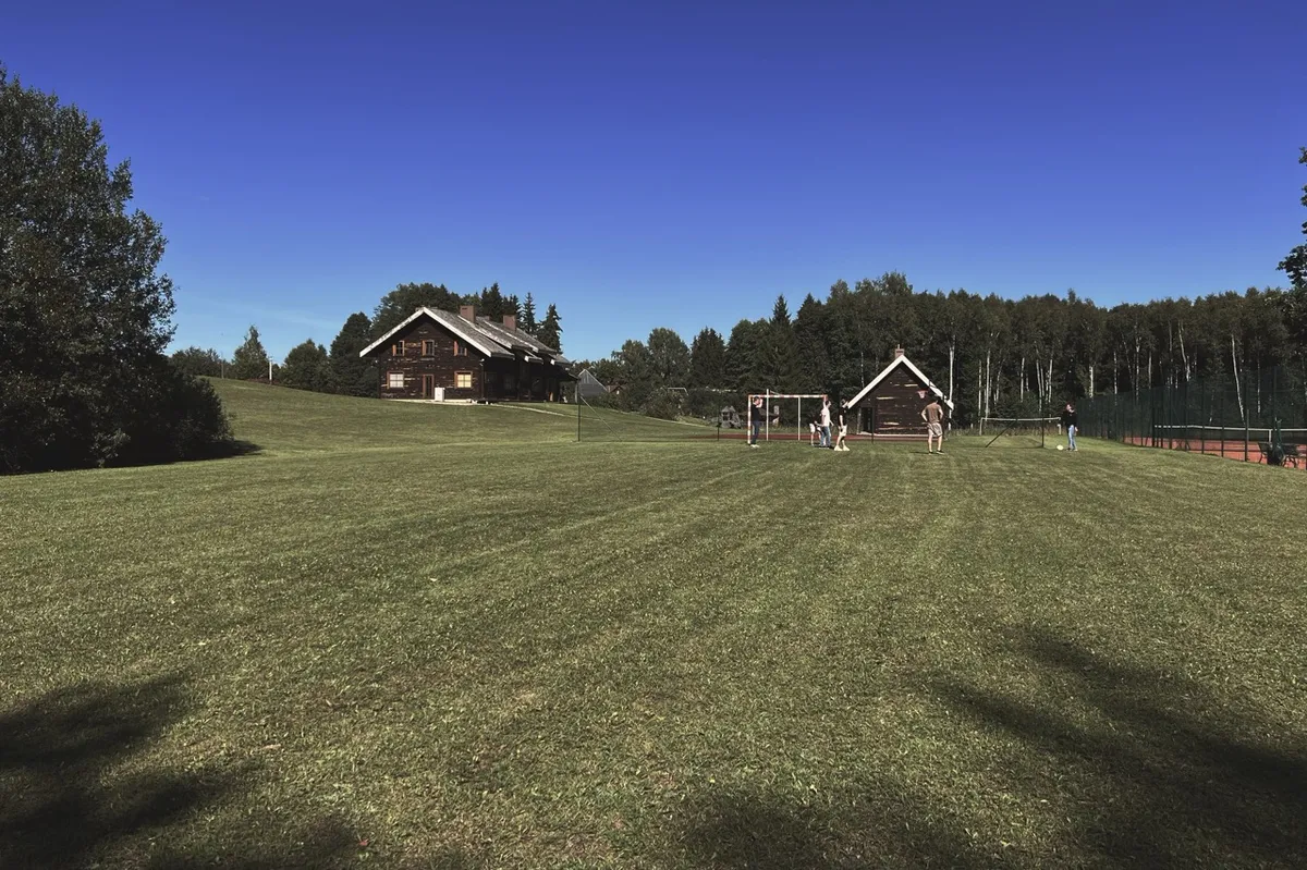 Wooden houses stand on a lush green field, surrounded by trees. A group of people plays volleyball in the open, under a clear blue sky.