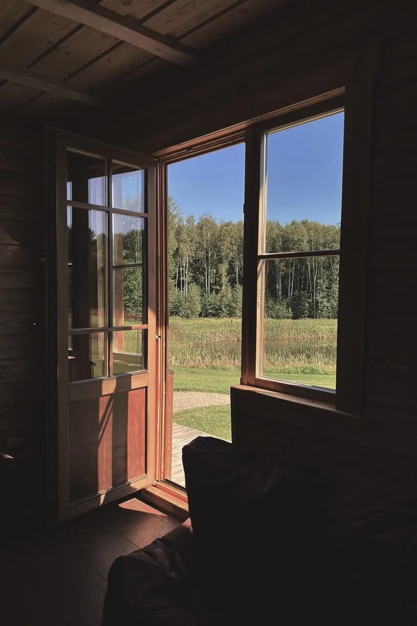 Sunlight streams through an open wooden door, revealing a view of green fields and a tree line under a clear blue sky, inside a rustic cabin.