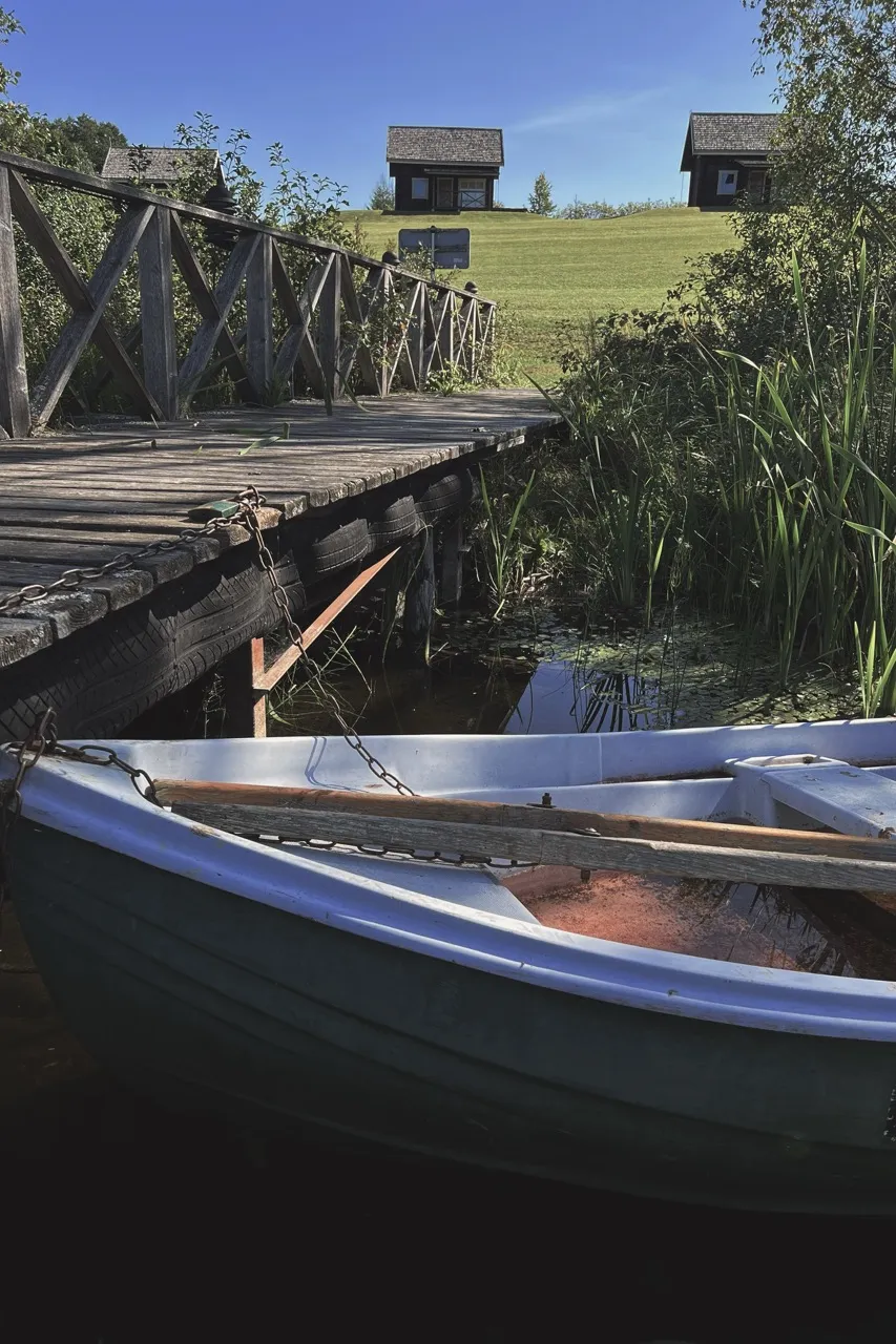 A small rowboat is tethered to a wooden dock amid lush greenery, with two wooden cabins on a grassy hill under a clear blue sky.