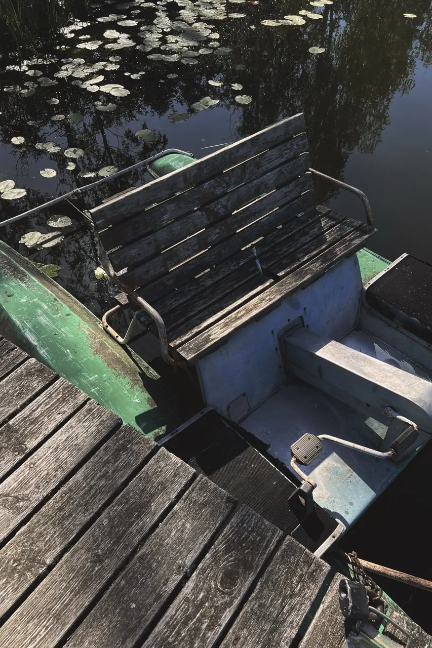 Wooden bench on a worn, green paddleboat docked beside a wooden pier, surrounded by lily pads floating on a calm, reflective water surface.