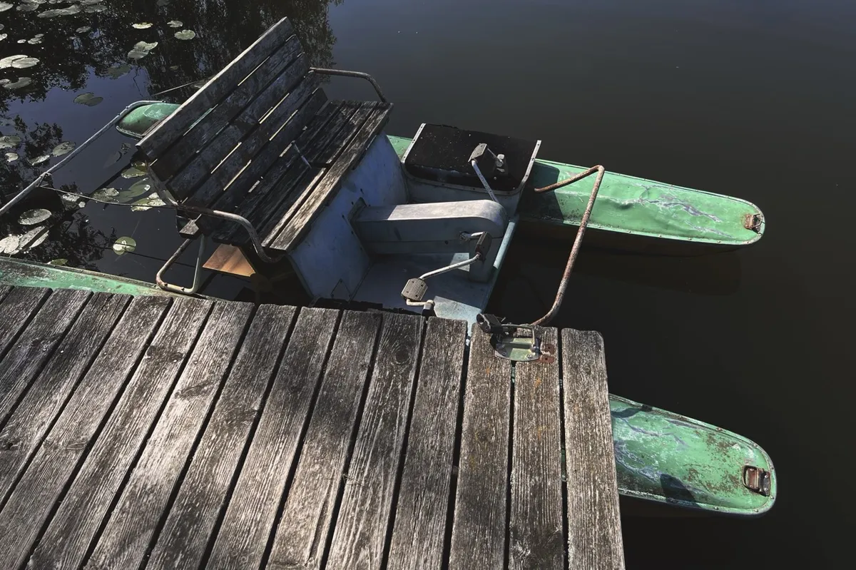 Weathered pedal boat with dual green pontoons, docked beside a wooden pier, floats on calm water partially covered with lily pads.