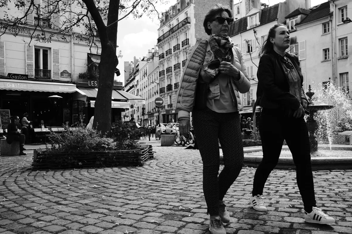 Two women walk on a cobblestone street past a fountain, surrounded by historic buildings and a café called "Gaston" in a lively European square.