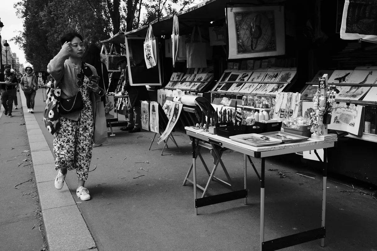 A person walks past a street art market, examining stalls displaying paintings, prints, and souvenirs. Trees line the pedestrian pathway, providing a casual, outdoor setting.