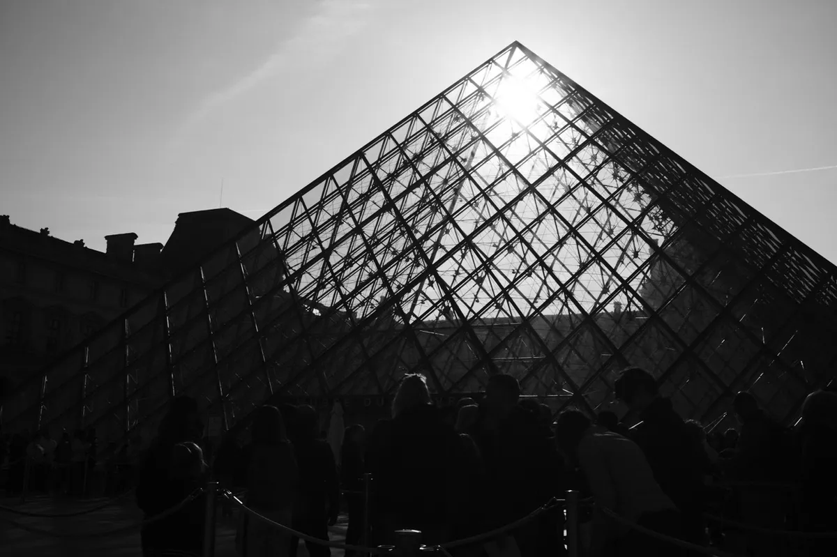 Glass pyramid reflecting sunlight with silhouetted people standing in front, set against a clear sky and surrounding historic buildings.