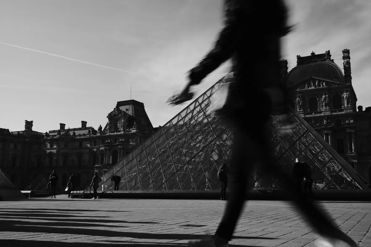Silhouette of a person walking across a plaza with a large glass pyramid and historic buildings in the background, under a clear sky.