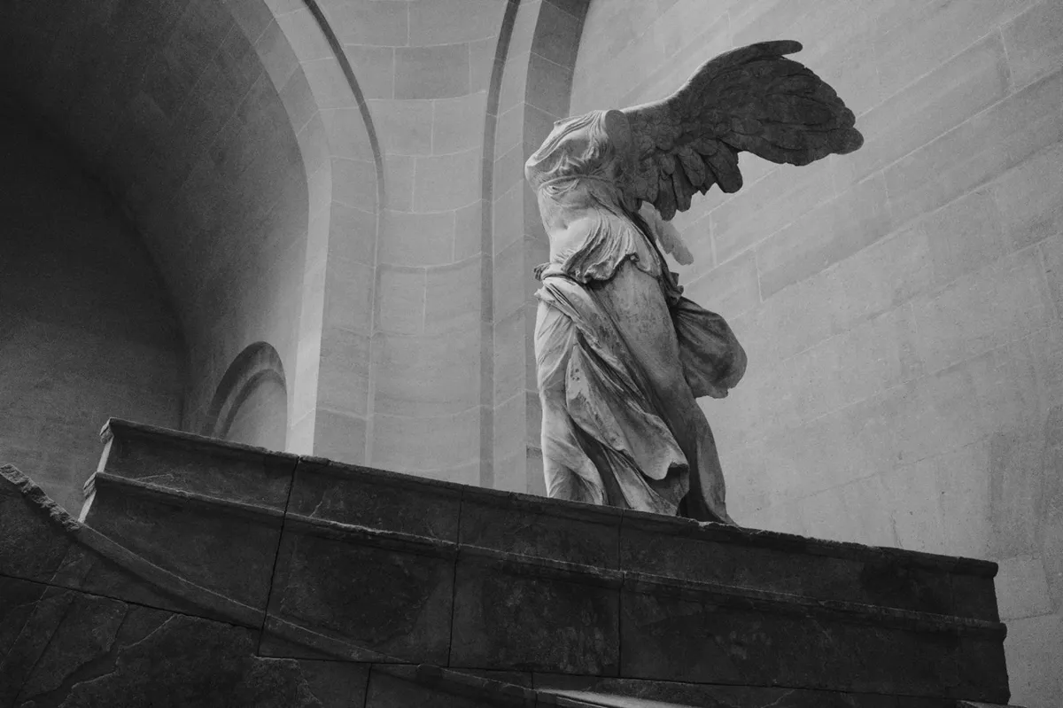 A headless marble statue with large wings stands gracefully poised, depicting movement, set atop a stone pedestal in a grand, arched architectural interior.
