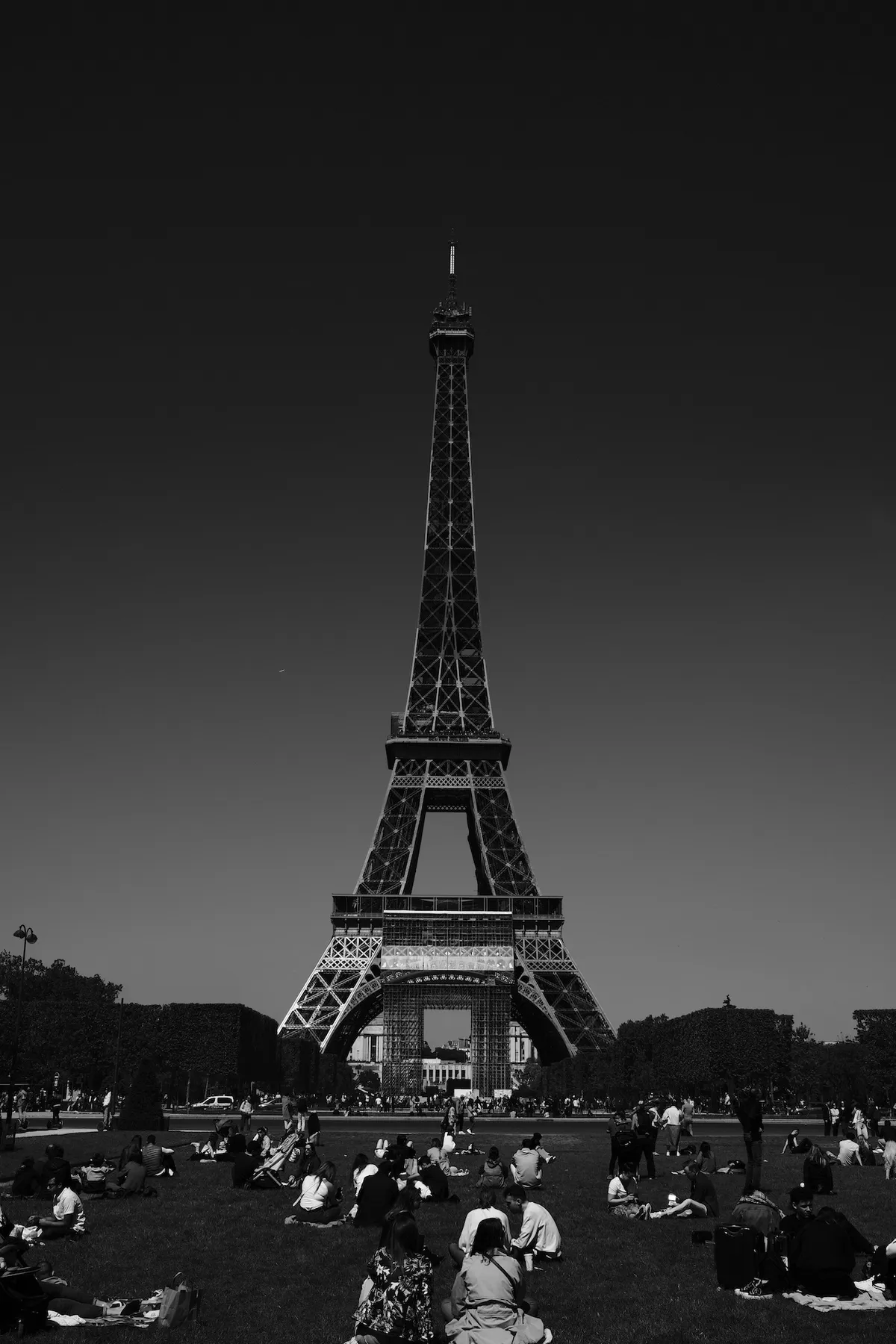 Eiffel Tower stands towering; people are seated, relaxing on grass in front. Bright sky creates a serene backdrop, trees framing the scene.