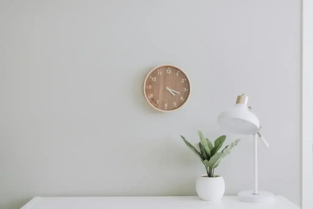 A simple desk with a simple plant, a simple lamp, and a simple clock above it.