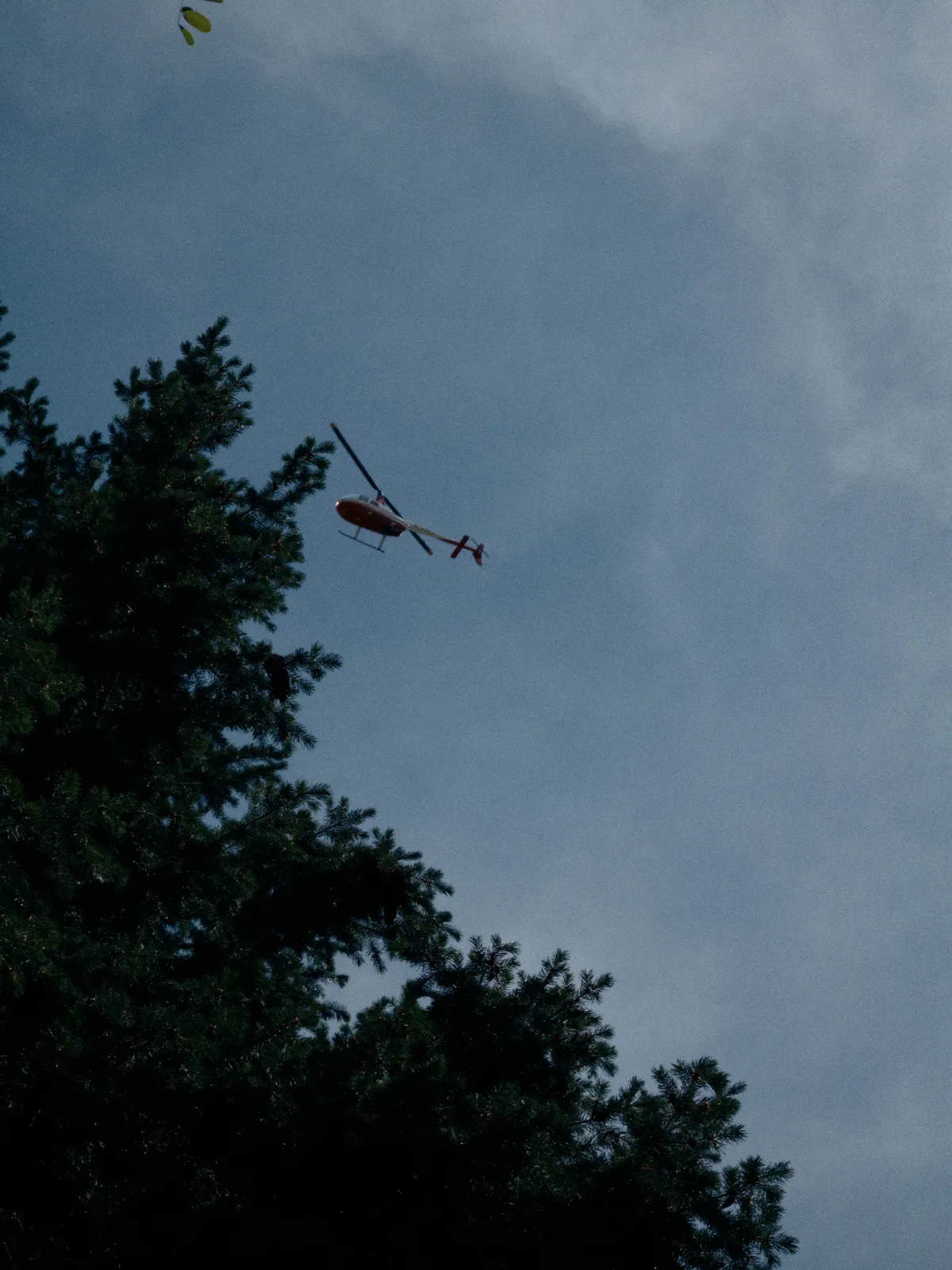 A dim, grainy shot looking up into the lightly clouded, blue sky, with dark, underexposed, green trees in the foreground. There is a red and white fire or rescue helicopter in the middle of the frame.