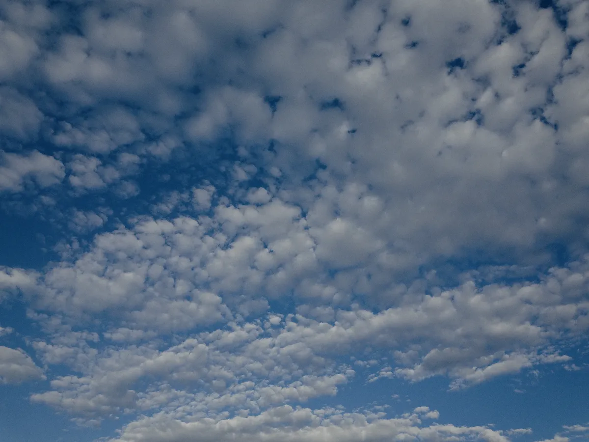 A grainy shot of a clouded sky. The sky is a deep, dreamlike blue, and the clouds are broken up into puffy chunks across the sky. The scene looks expansive and large.