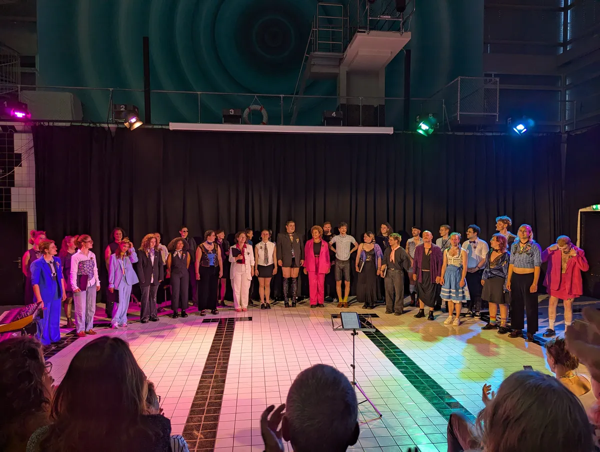 The choir singers stand in a semi-circle facing the audience, addressing their applause. They are all dressed fabulously.