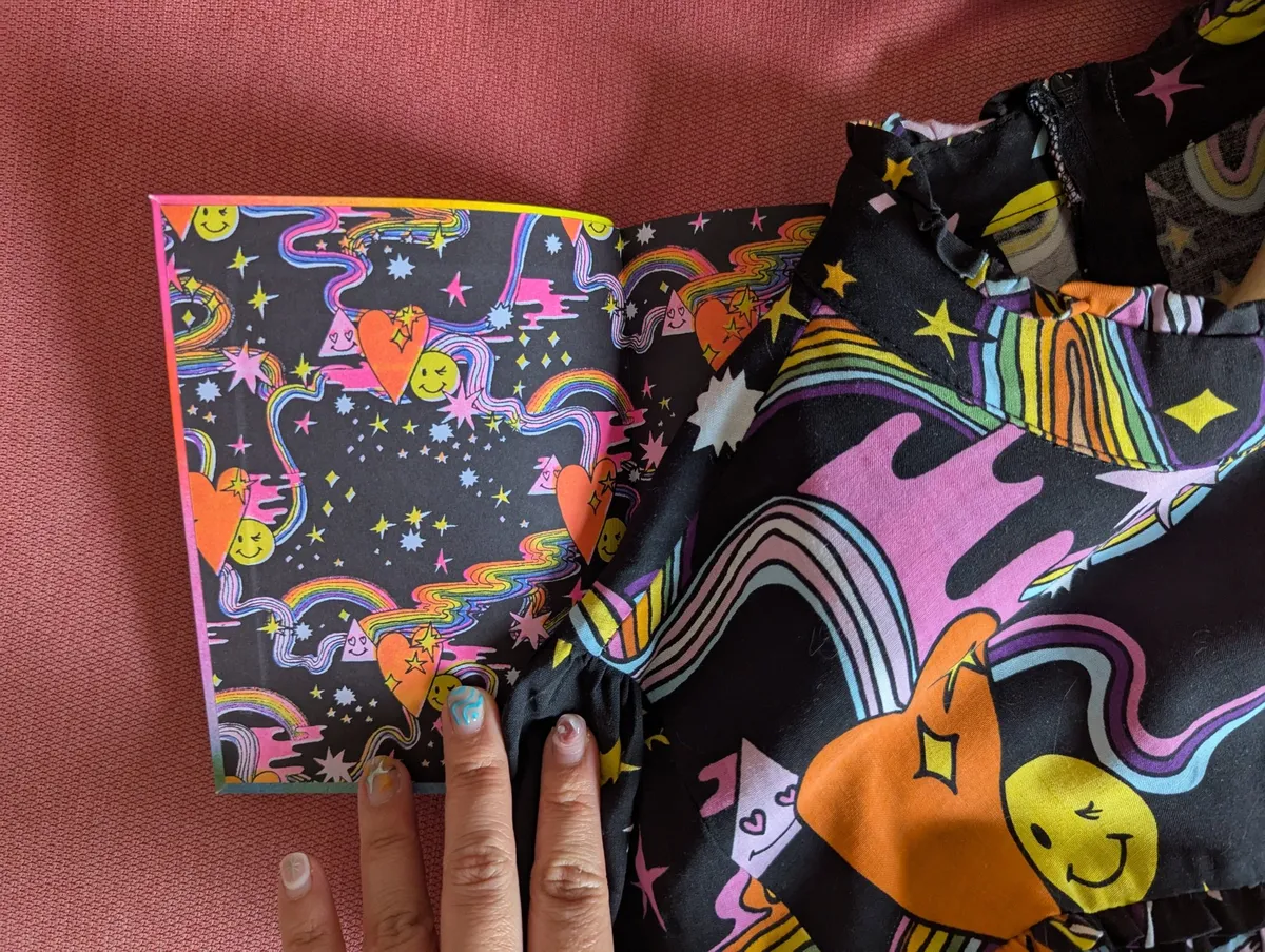 The opening spread of the guidebook depecting a psychedelic pattern with rainbows, hearts, and happy faces. Lying on top is a dress with the same pattern.