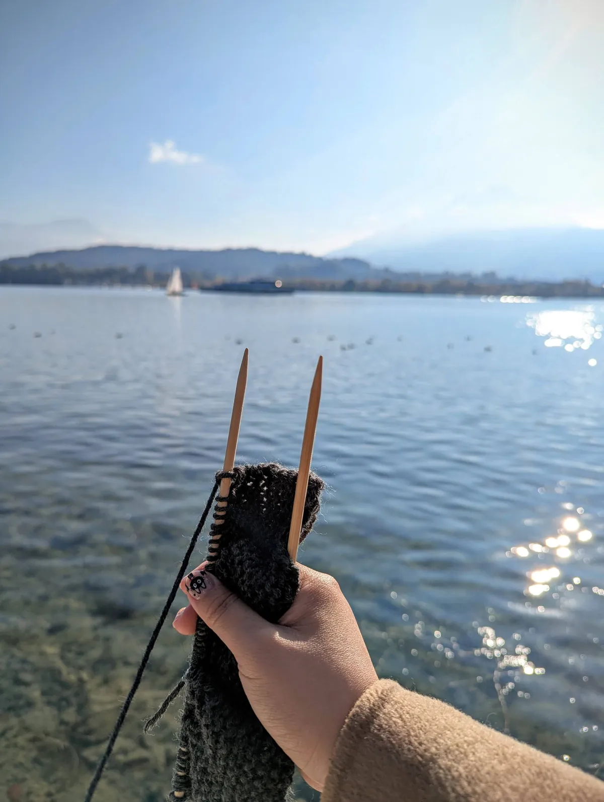 Peige holding the beginnings of a knitted gray scarf against a lakeside view.