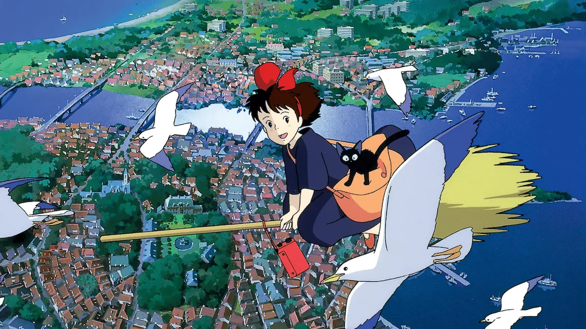 Kiki flying over a European-looking town. She is surrounded by seagulls and Jiji is perched on her orange bag, looking alarmed.