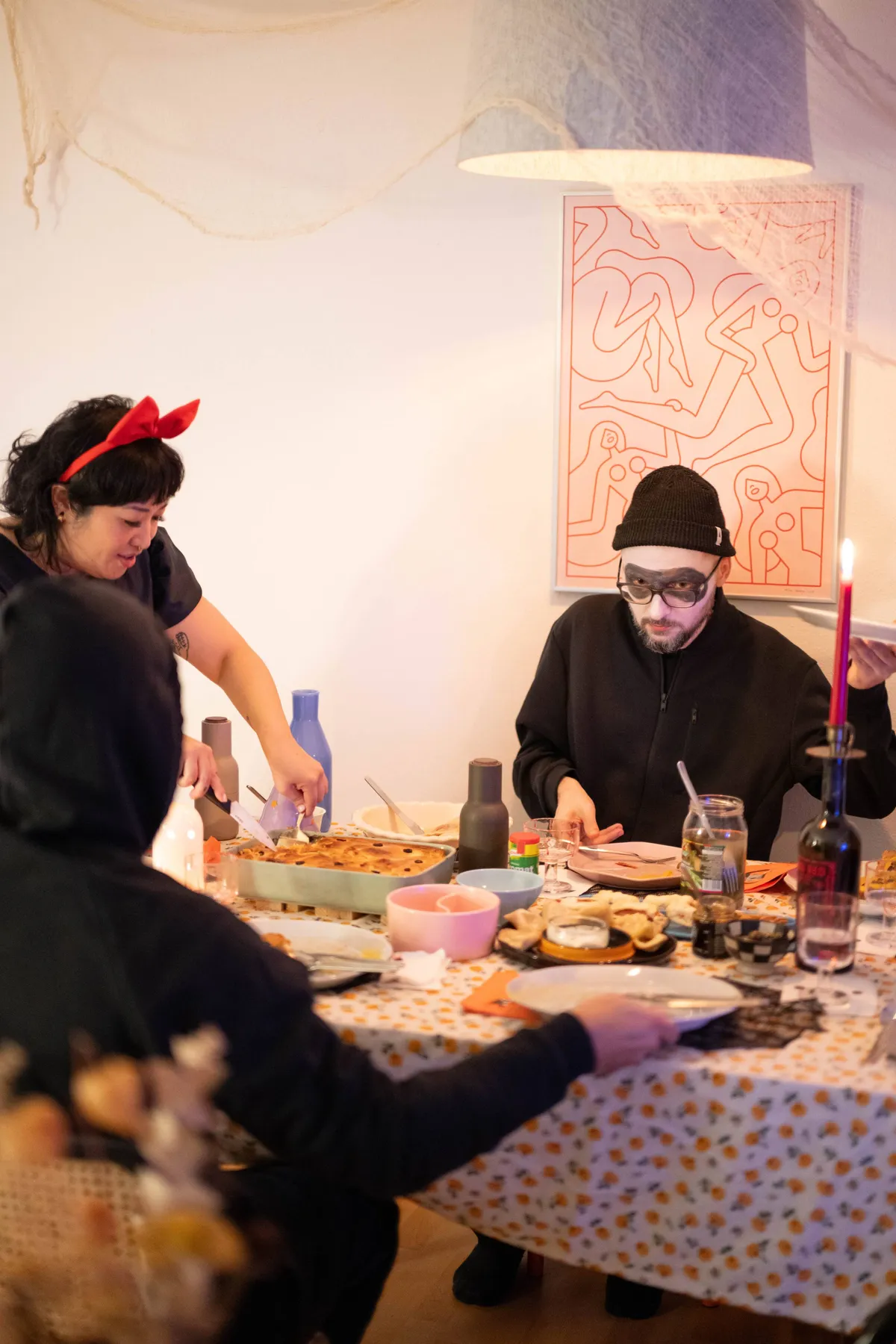 Peige serving the guests pie. Daniel is sitting facing the camera, his face painted white with a black "eye mask" painted around his eyes.