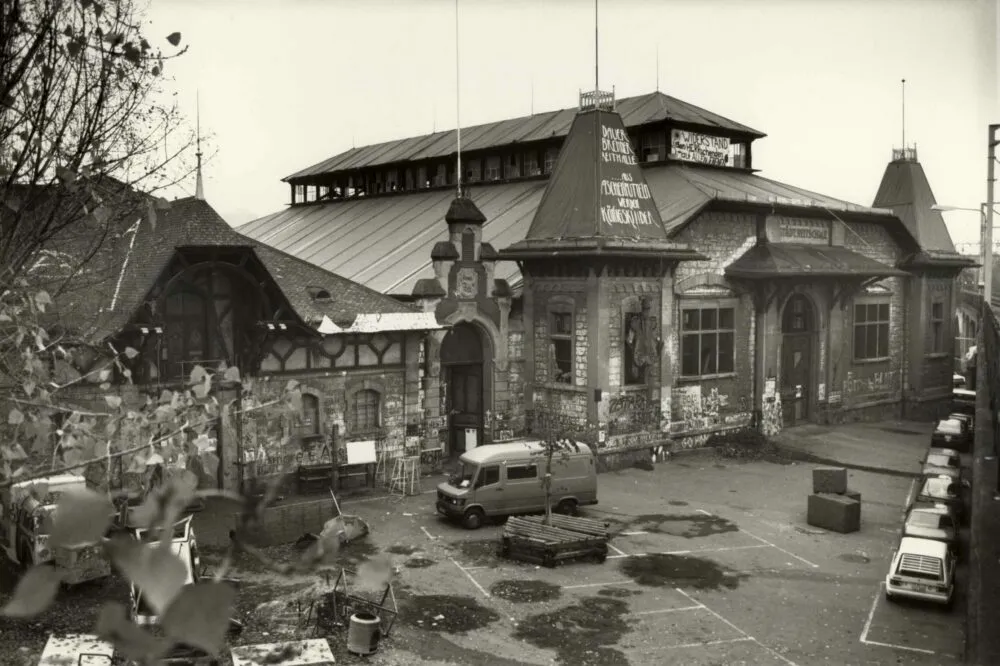 A black and white photo of a large building with some graffiti and cars parked outside.