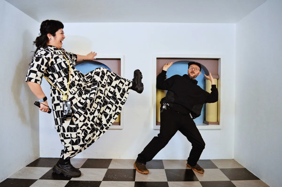 Peige and Daniel posing in an optical illusion room, where Peige is tall and Daniel is small. Peige pretends to kick Daniel while Daniel leans back with his arms in the air.