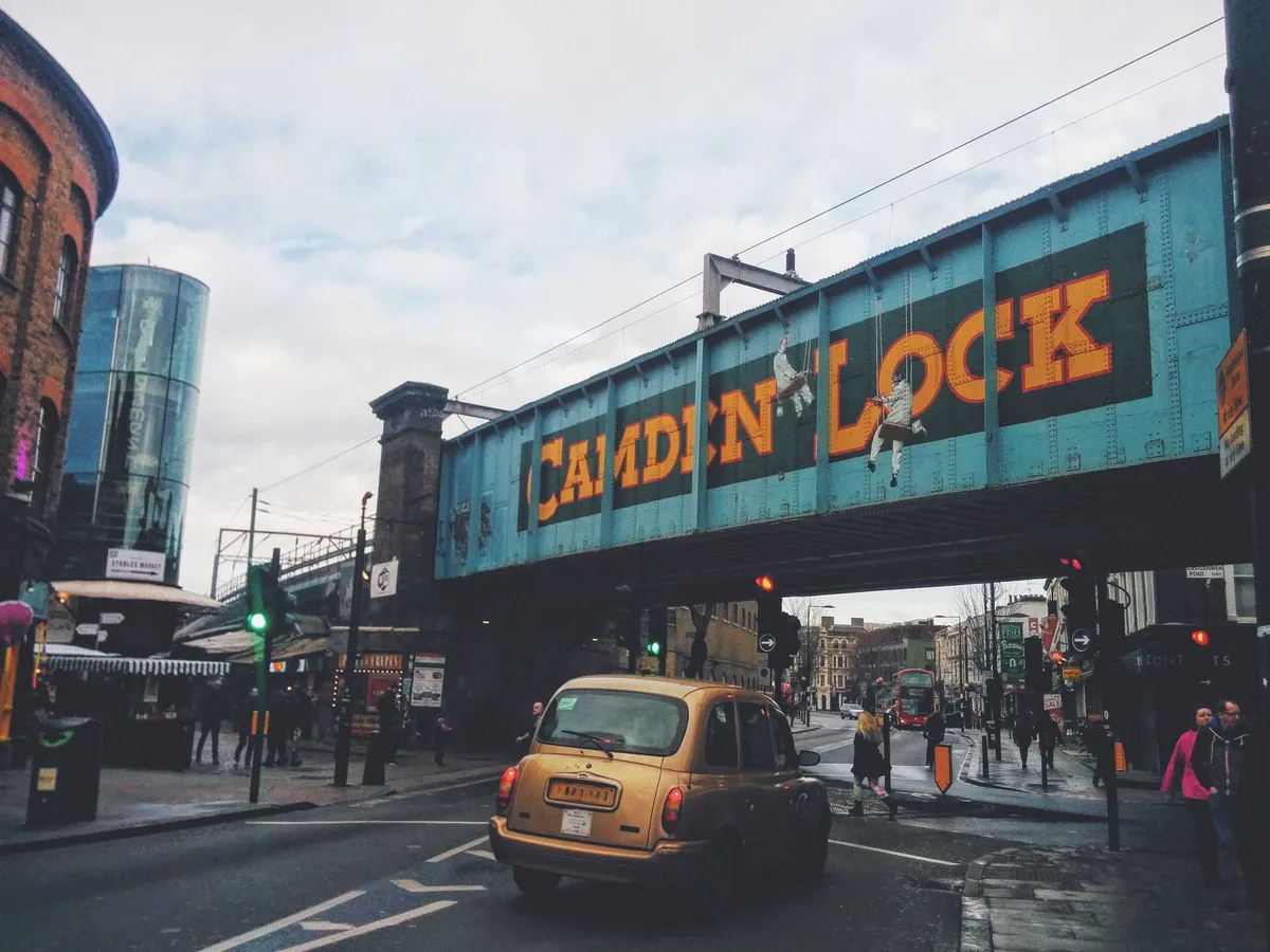 A golden taxi stopped at an intersection. Above is a giant bridge with "Camden Lock" painted on it.