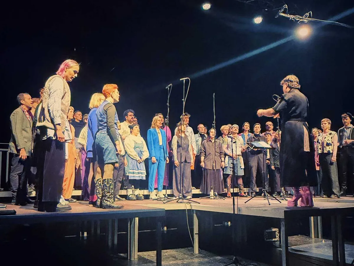 A group of about 50 people standing in a semi-circle, singing together. The conductor is wearing pink, 10-inch platform boots.