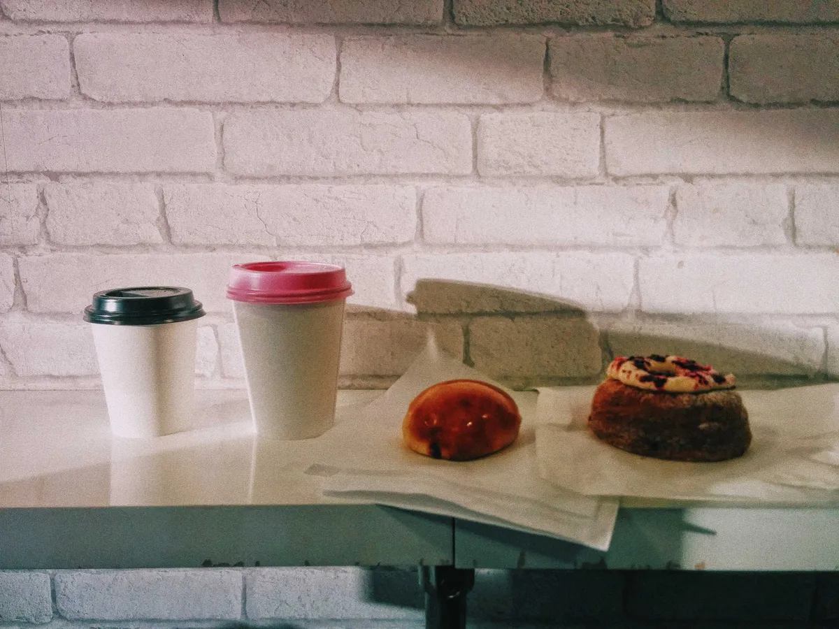 Two coffees in a takeout cup (one with a black lid, one with a pink lid), one glazed Berliner and a fancy-looking cronut.