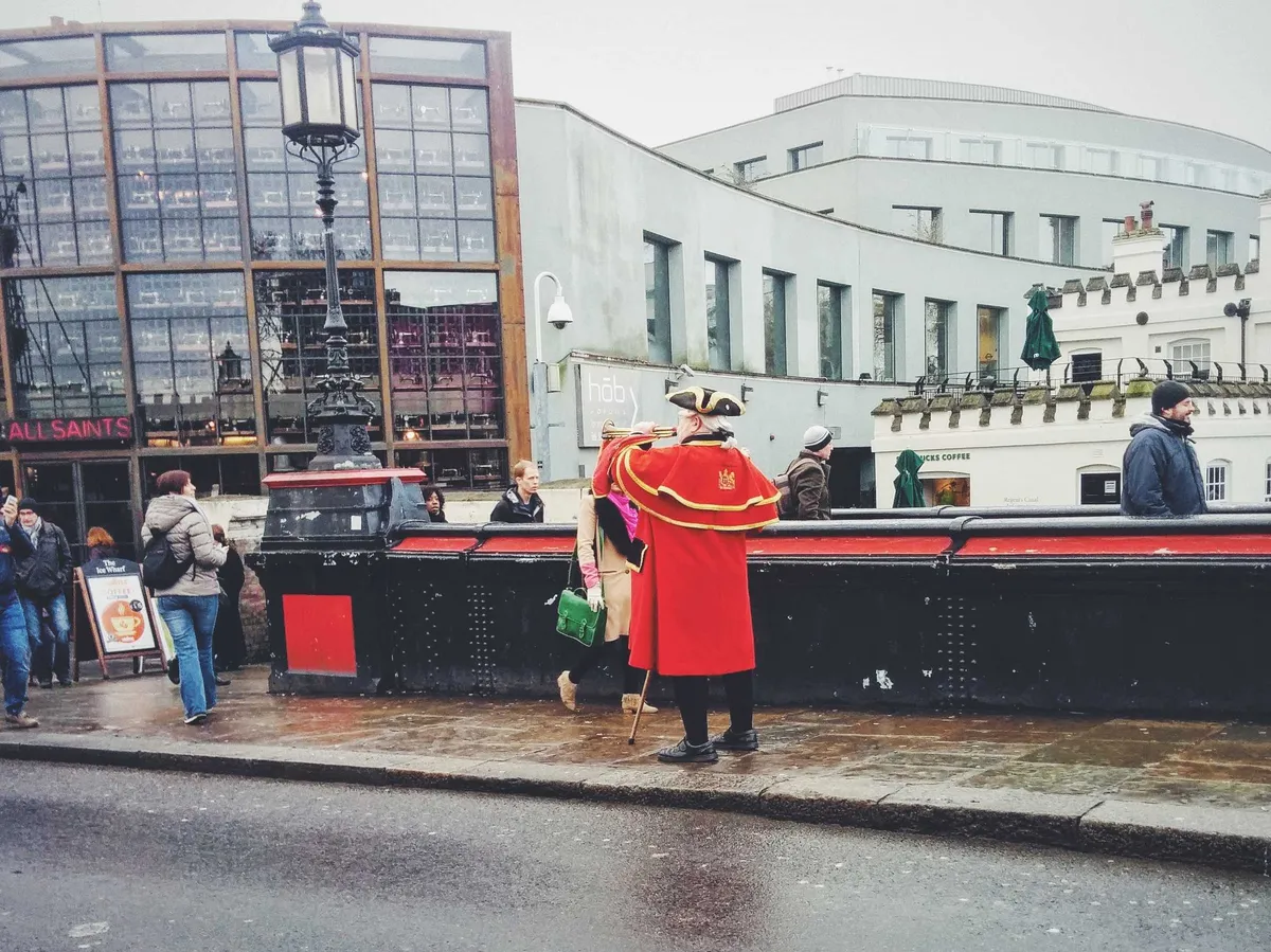 A man wearing a traditional British costume blowing a trumpet in the middle of a street.