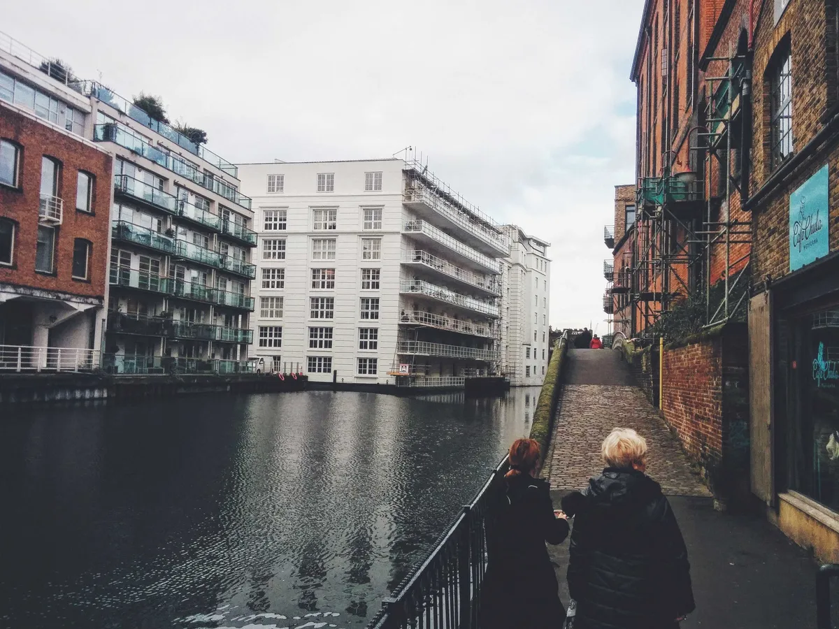 Two people walk alongside the canal. Modern buildings are on the left of the canal, while the right side has brick-and-mortar buildings.
