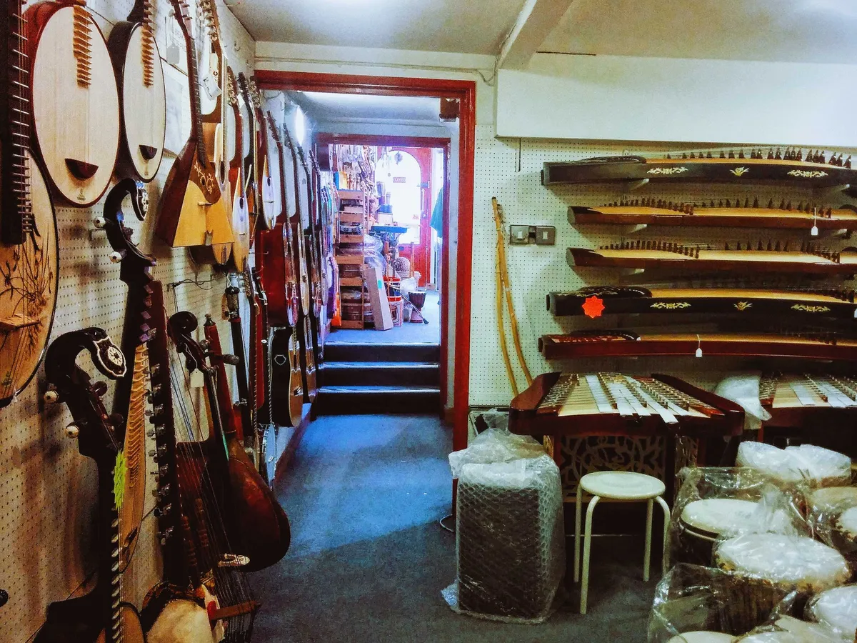 A wall filled with mandolins and guitars. Another wall contains rows of guzheng. Towards the floor are drums wrapped in plastic.