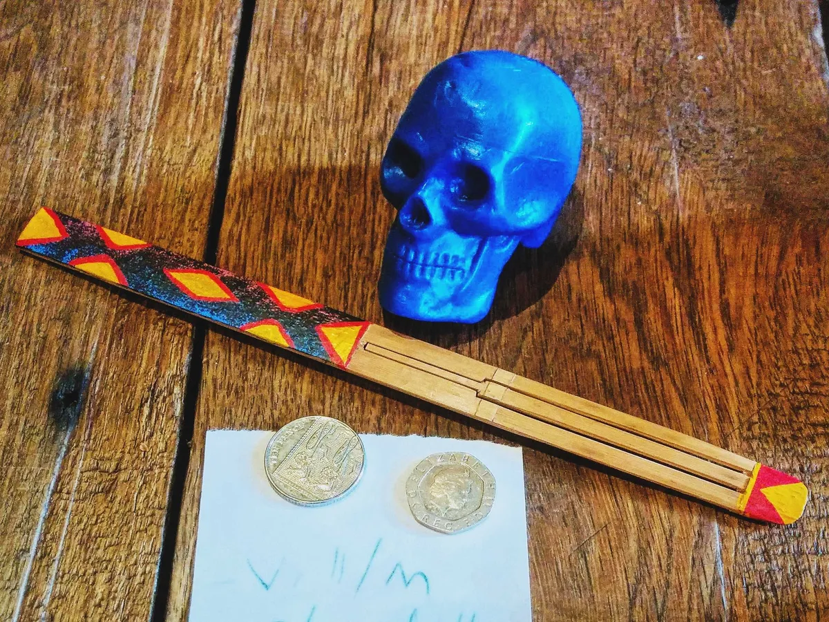 A blue skull next to a colorful bamboo stick. There are two coins on the table on top of a white piece of paper with Peige's lunch order written in colored pencil.