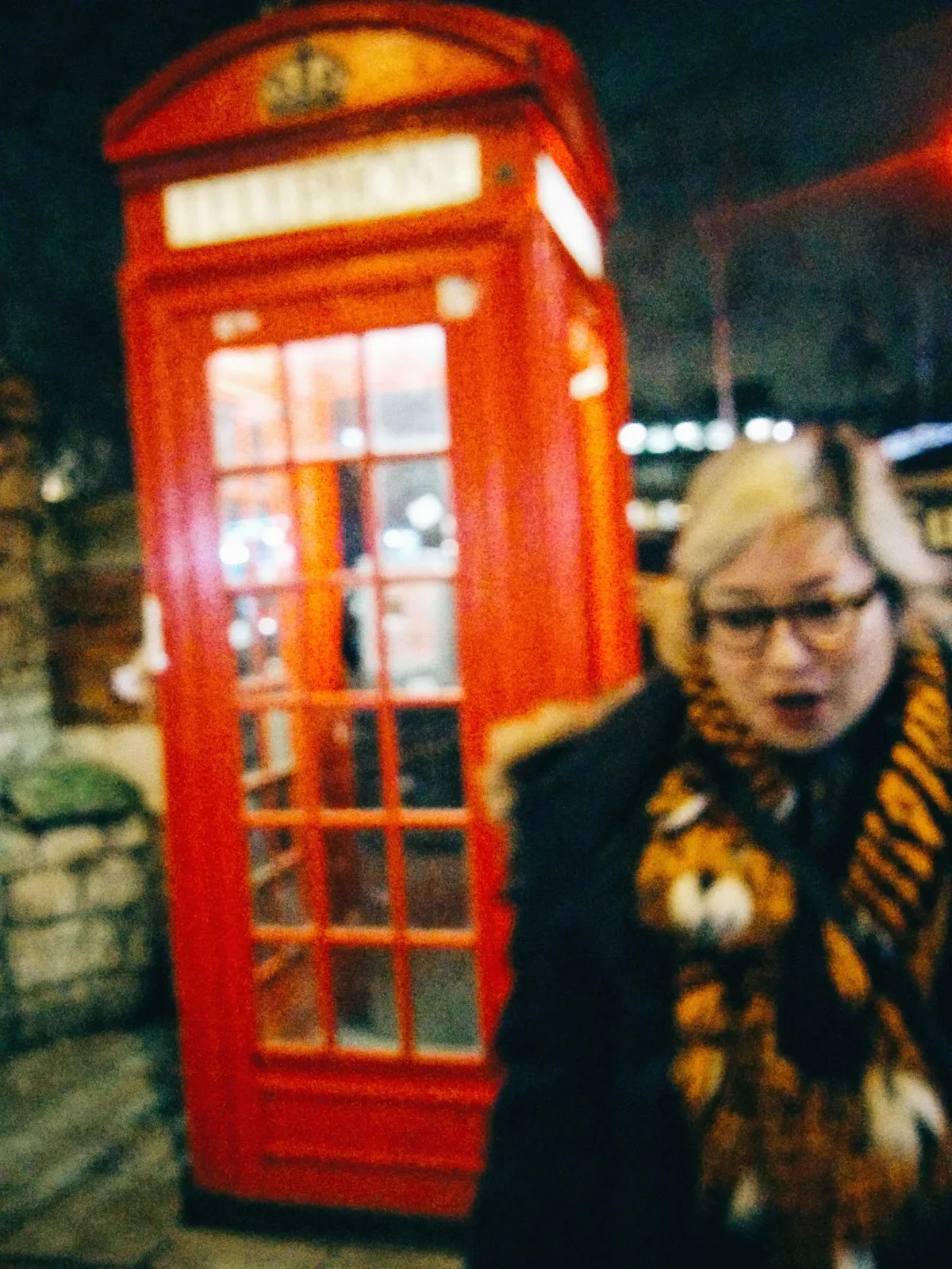 Peige walking away from the empty red phone booth. Their mouth is slightly open, as if they are talking.