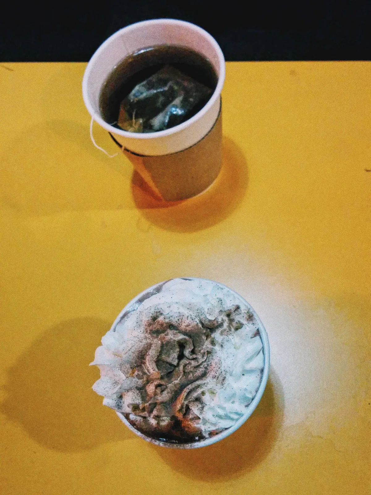 A cup of tea and a whipped drink on top of a bright yellow table.