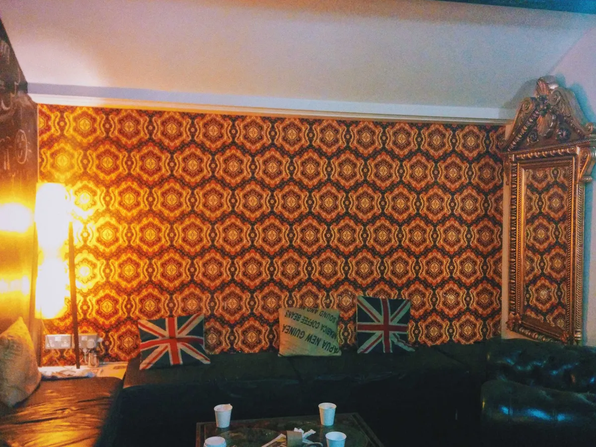 A sitting area with loudly patterned orange and brown wallpaper. There are black leather couches and pillows with the Union Jack printed on them.