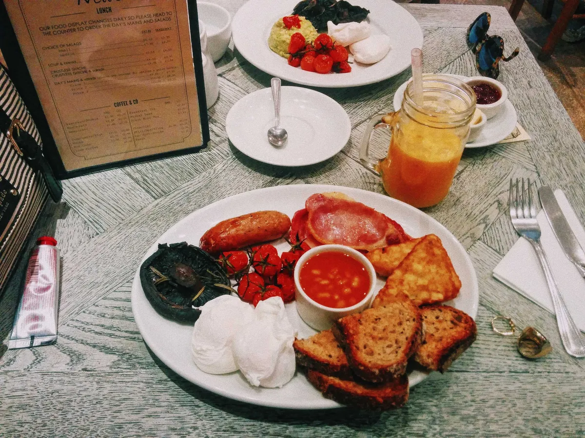 A plate containing toast, hash browns, bacon, a mushroom, small tomatoes, sausages, and two poached eggs.