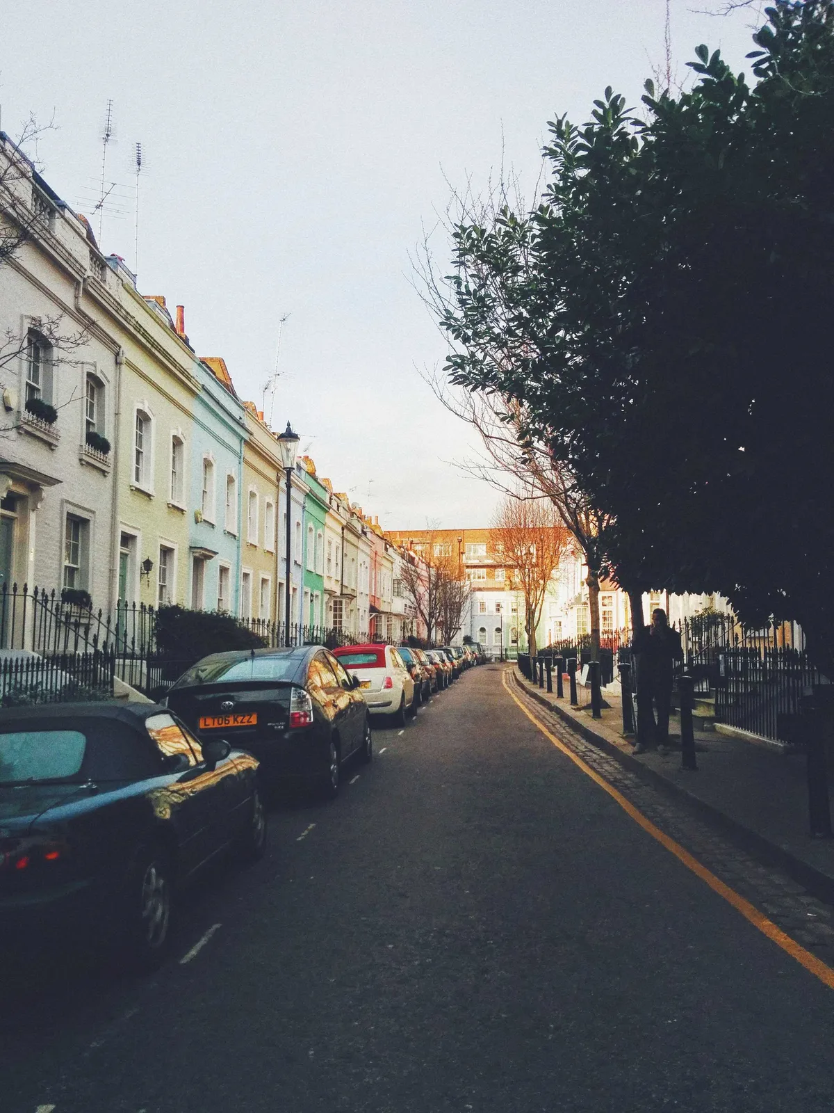 Street with colorful buildings and parked cars on one side, and shaded trees on the other.