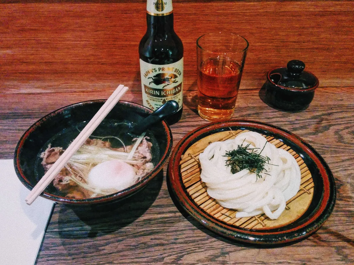 A bowl of soup with beef slices next to a bamboo plate of udon, accompanied by a Kirin Ichiban beer.