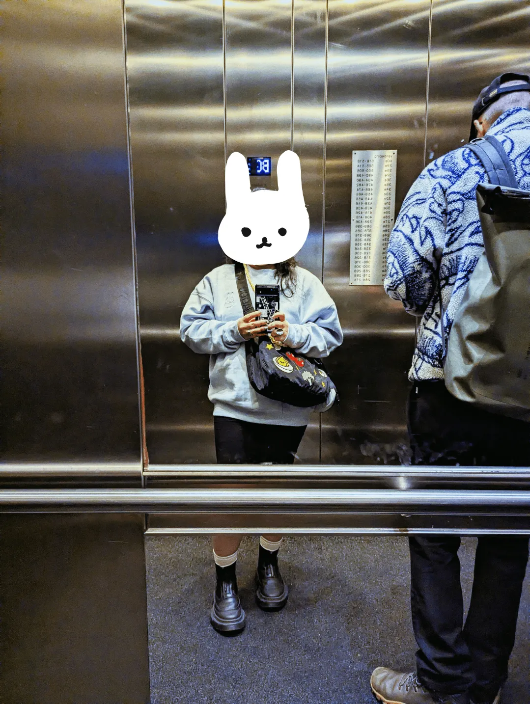 Peige standing in front of the mirror inside of the elevator. They are wearing a light blue sweater, black shorts, socks, and black boots. Daniel can be seen leaning against the mirror.
