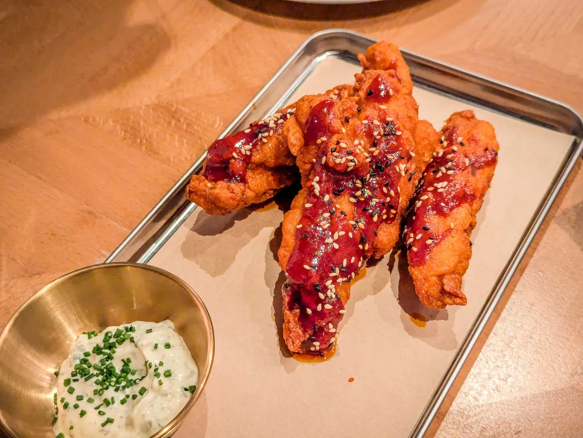 A small pile of chicken tenders smothered in red sauce. A tiny container of white sauced sprinkled with chives is on the side.