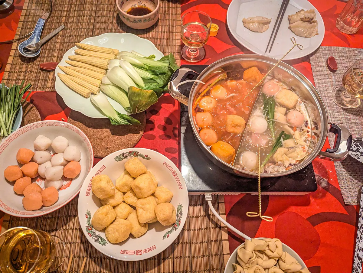 A colorful table spread of hot pot ingredients, such as fish balls and veggies, along with the hot pot containing two types of soups.