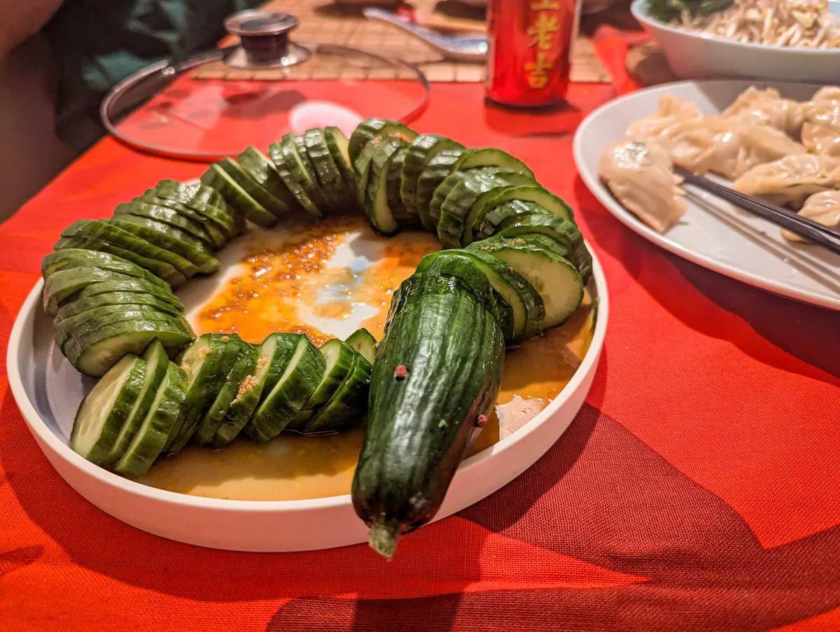 A plate containing a cucumber that has been sliced to look like a snake. The snake's "head" is hanging slightly off the plate, with it's "tongue" (the stem) sticking out.