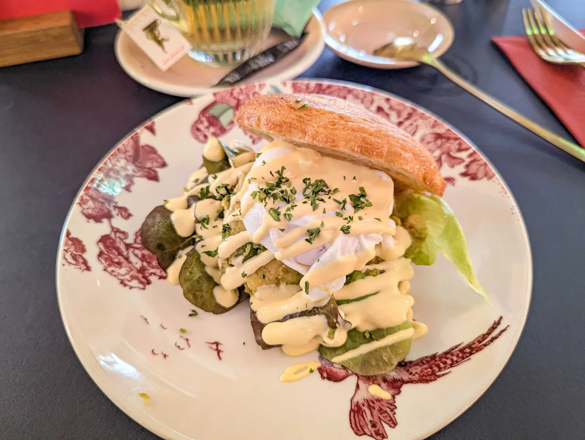 A croissant that has been filled with mashed avocado, salad, and a poached egg. It is smothered in hollandaise sauce.