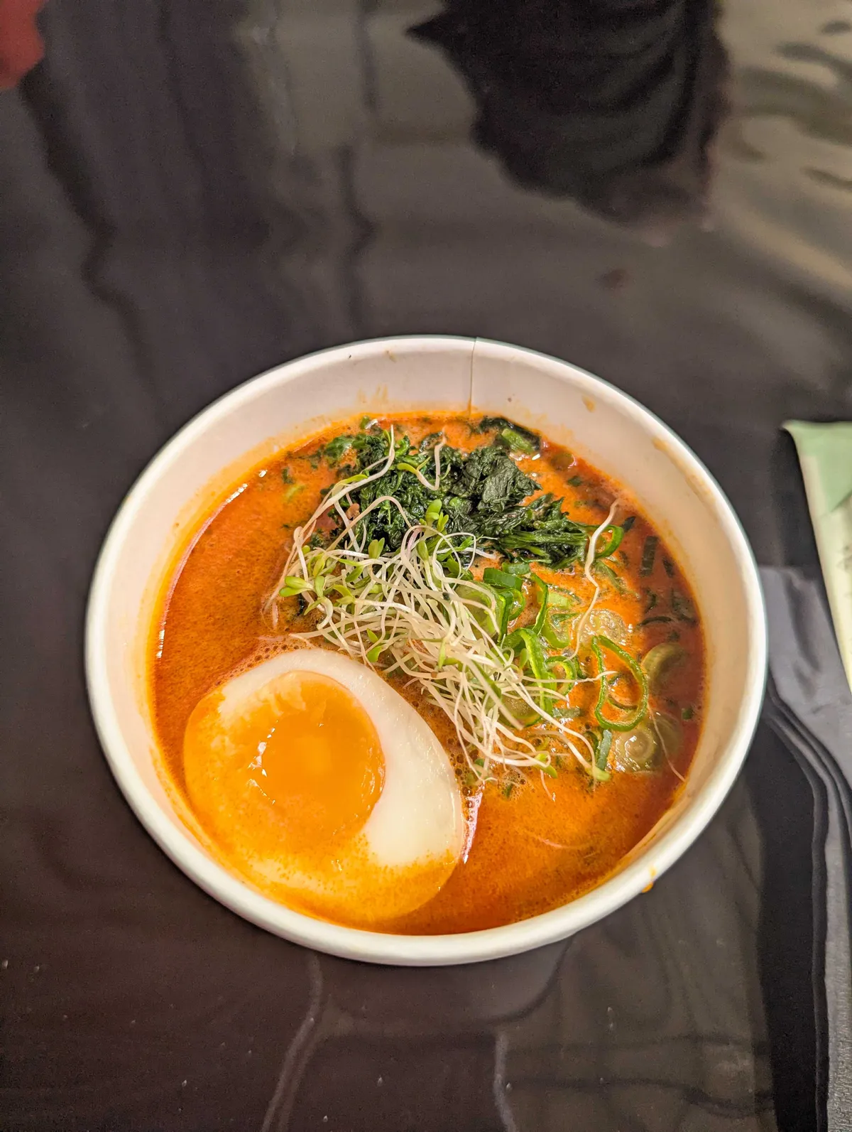 A bowl of spicy-looking soup filled with sprouts, seaweed, spring onions, and a seasoned soft-boiled egg.