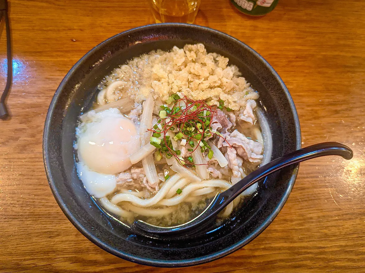 A delicious bowl of udon with an onsen egg, tempura bits, beef, spring onions, and onion sprouts.