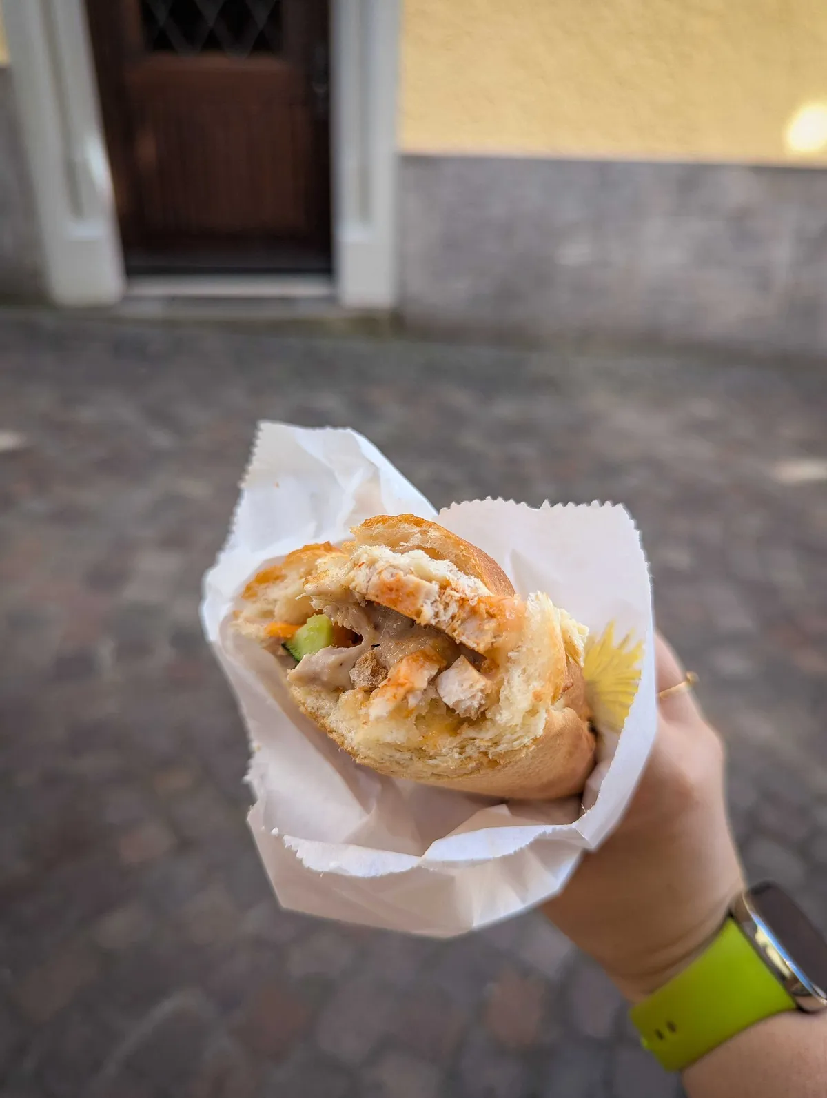Peige holds a bitten banh mi sandwich wrapped in white paper. Vietnamese sausage, cucumber, and sriracha are visible.