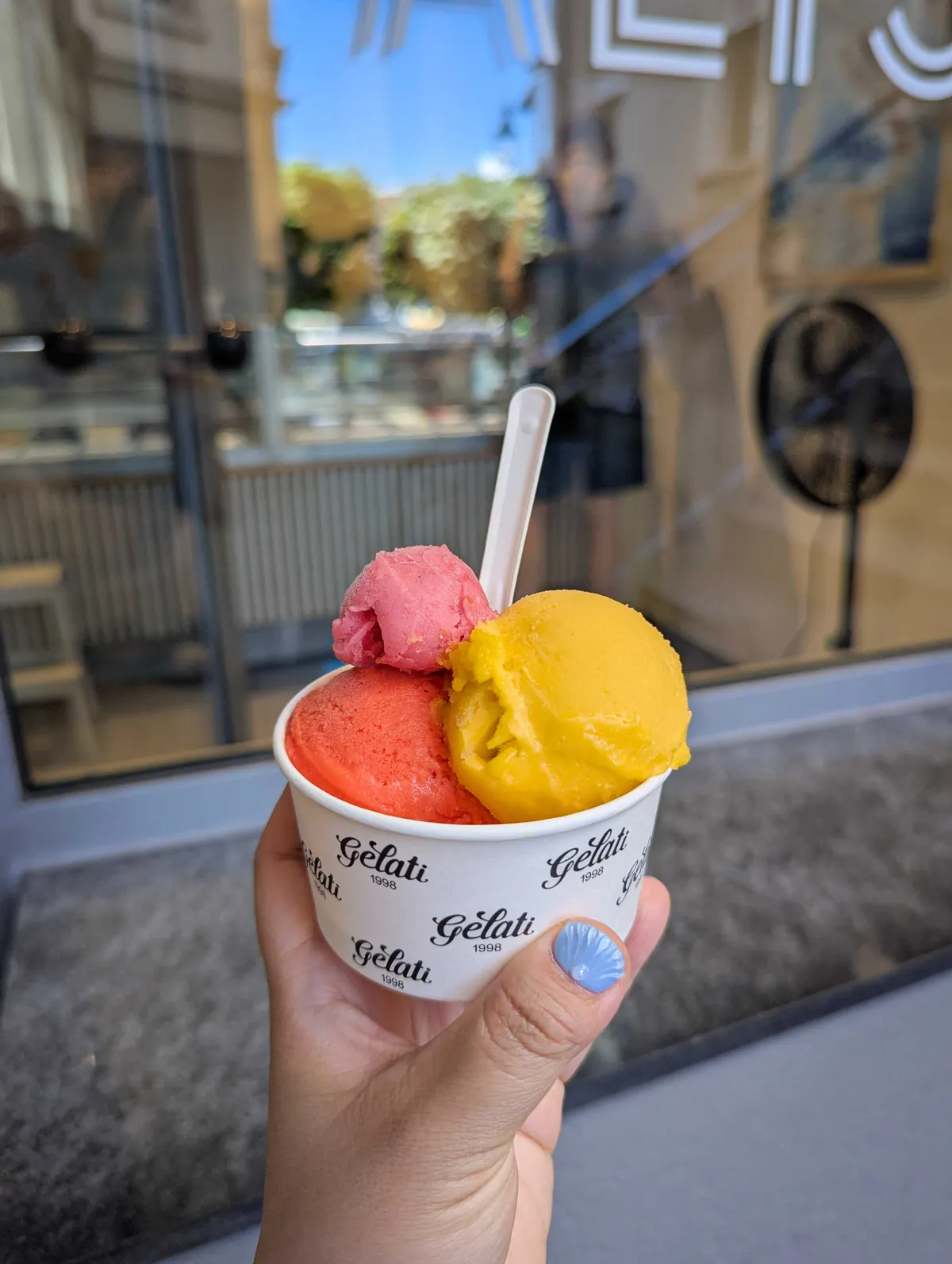 Peige holds a small cup containing three kinds of sorbets: A bright red-orange scoop, stacked on top with a golden yellow scoop, with a tiny pink scoop on top.