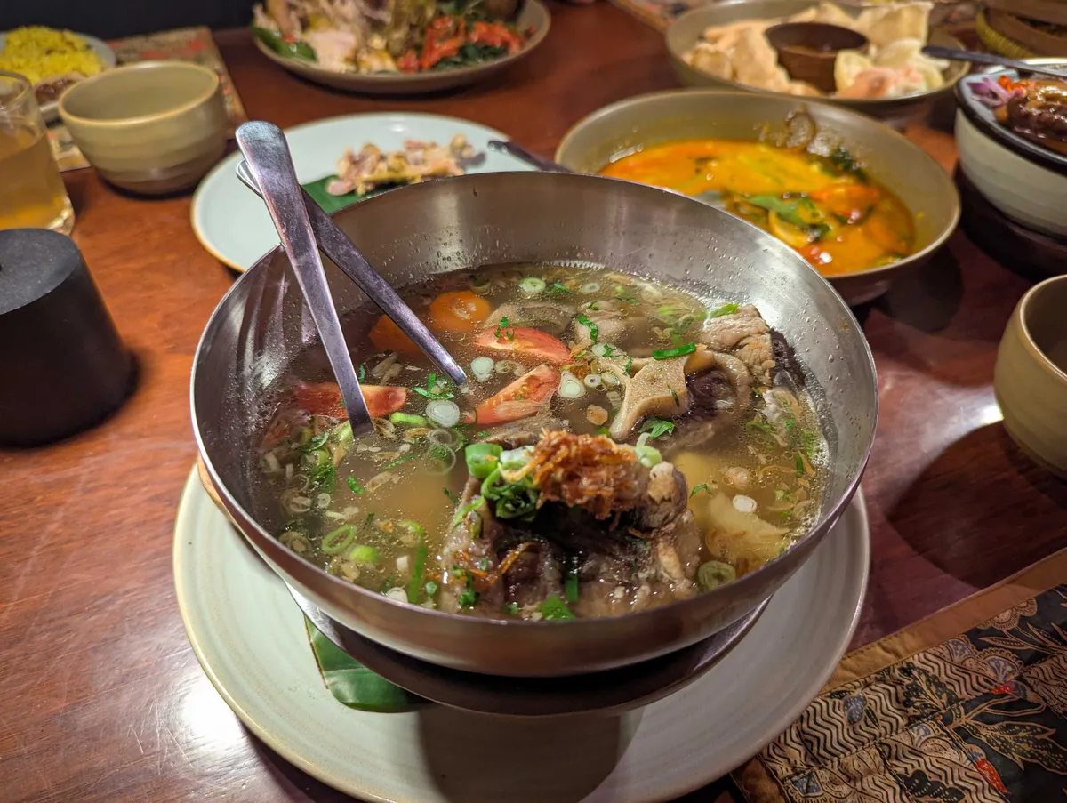 A large metal bowl filled with oxtail soup, sliced tomatoes, spring onions, and crispy fried onions.