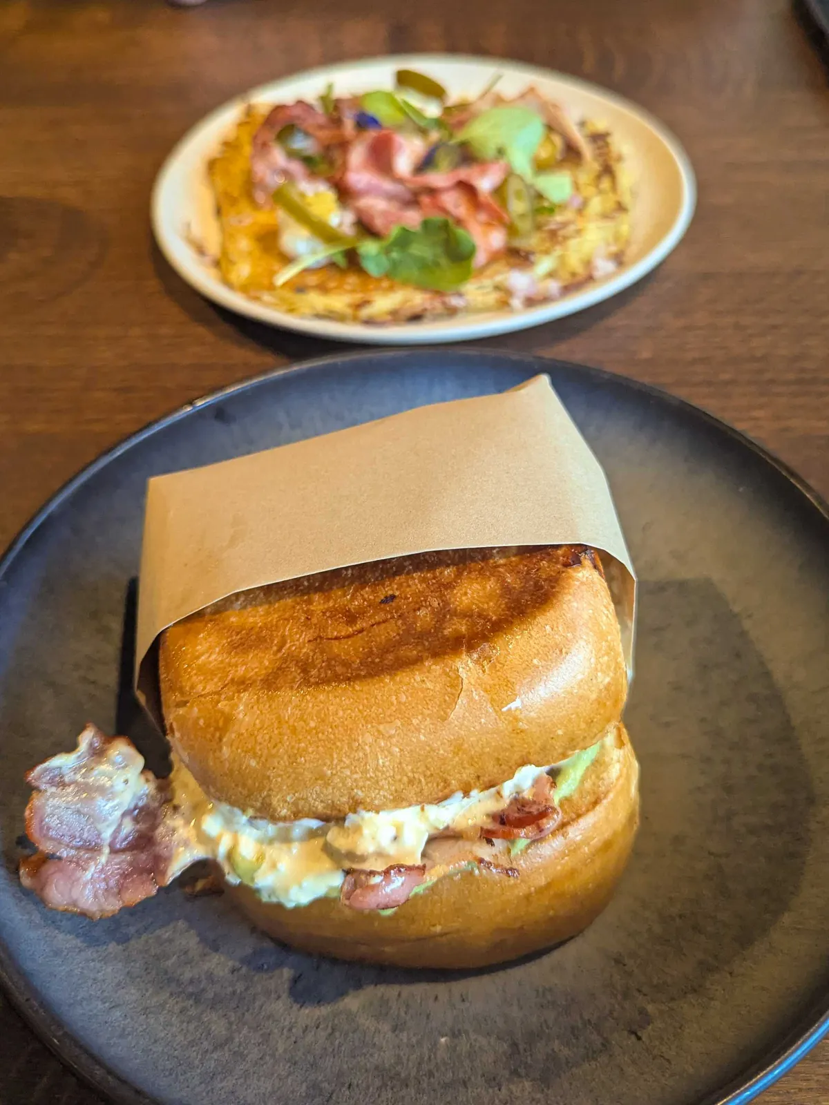 A thick, brioche sandwich filled avocado cream and bacon peeking out. Behind the sandwich is a plate of rösti.
