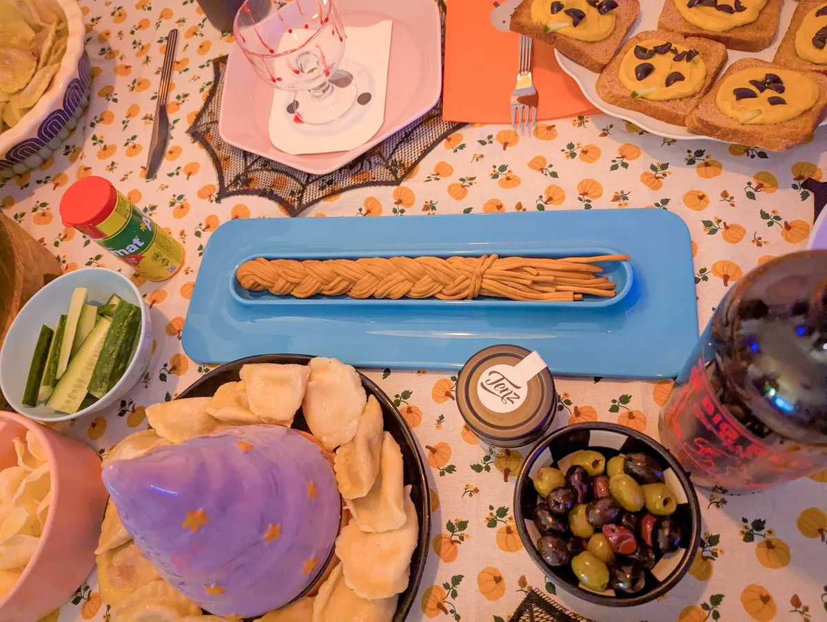 A colorful spread of Halloween-themed food. In the center is a roasted strips of cheese that have been braided to look like a broomstick. It is lying within a sky-blue colored plate.